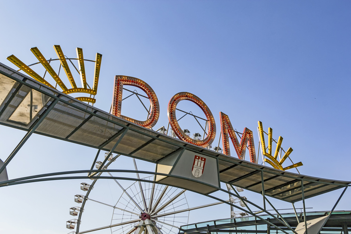 Zeichen über dem Eingang des Hamburger Doms: DOM in großen Buchstaben, dahinter steht das Riesenrad, blauer Himmel